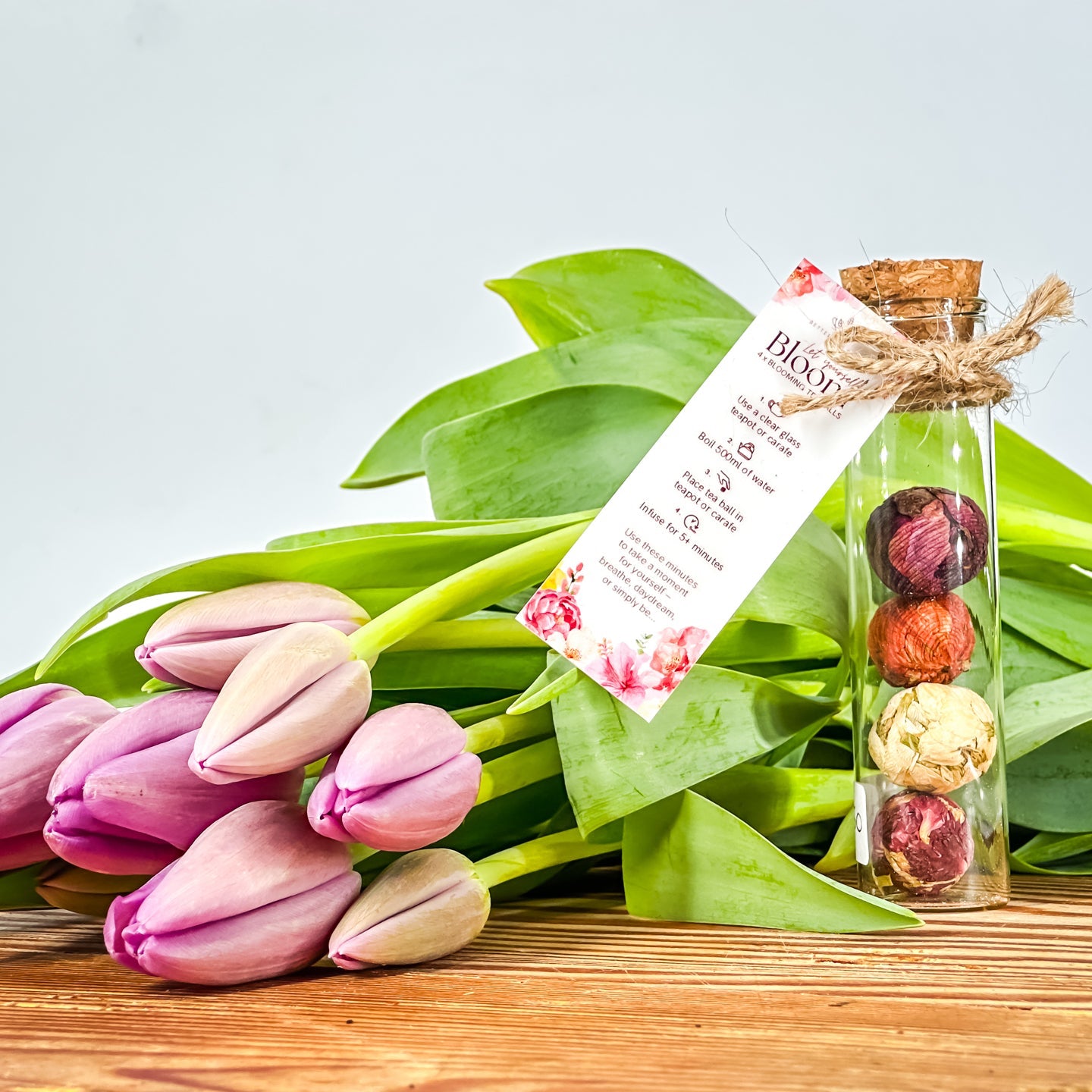floral tea balls in a glass tube with a clear mug with a bloomed flower tea steeping in hot water on a bread board