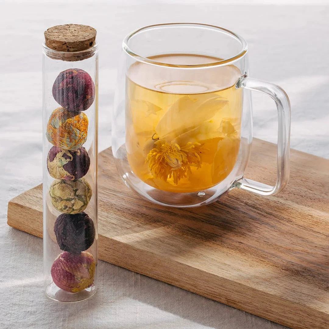 floral tea balls in a glass tube with a clear mug with a bloomed flower tea steeping in hot water on a bread board