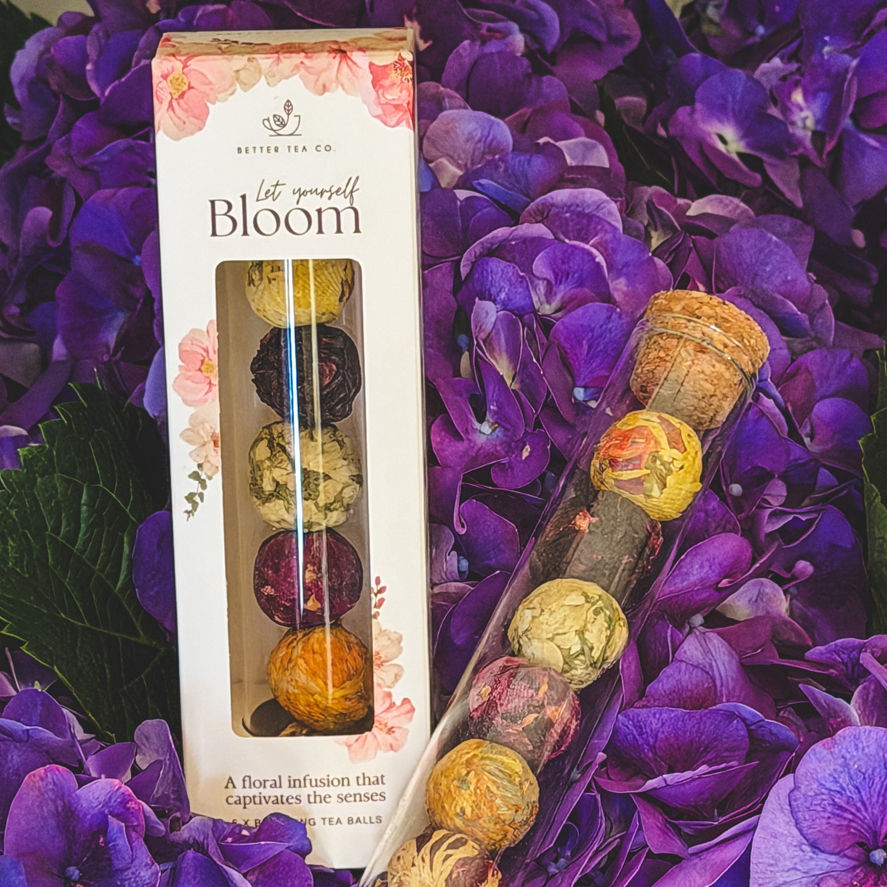 floral tea balls in a glass tube with a clear mug with a bloomed flower tea steeping in hot water on a bread board