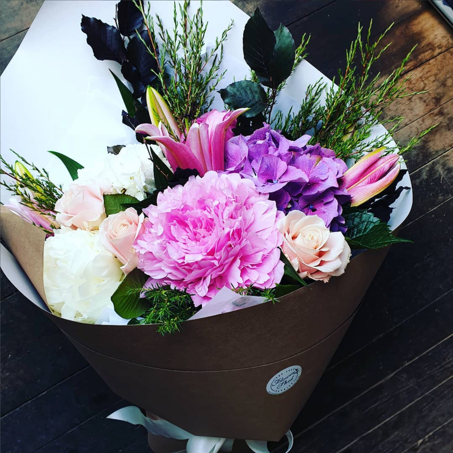 Bouquet of flowers with pink, purple, and white flowers in a brown paper wrap on a wooden surface.