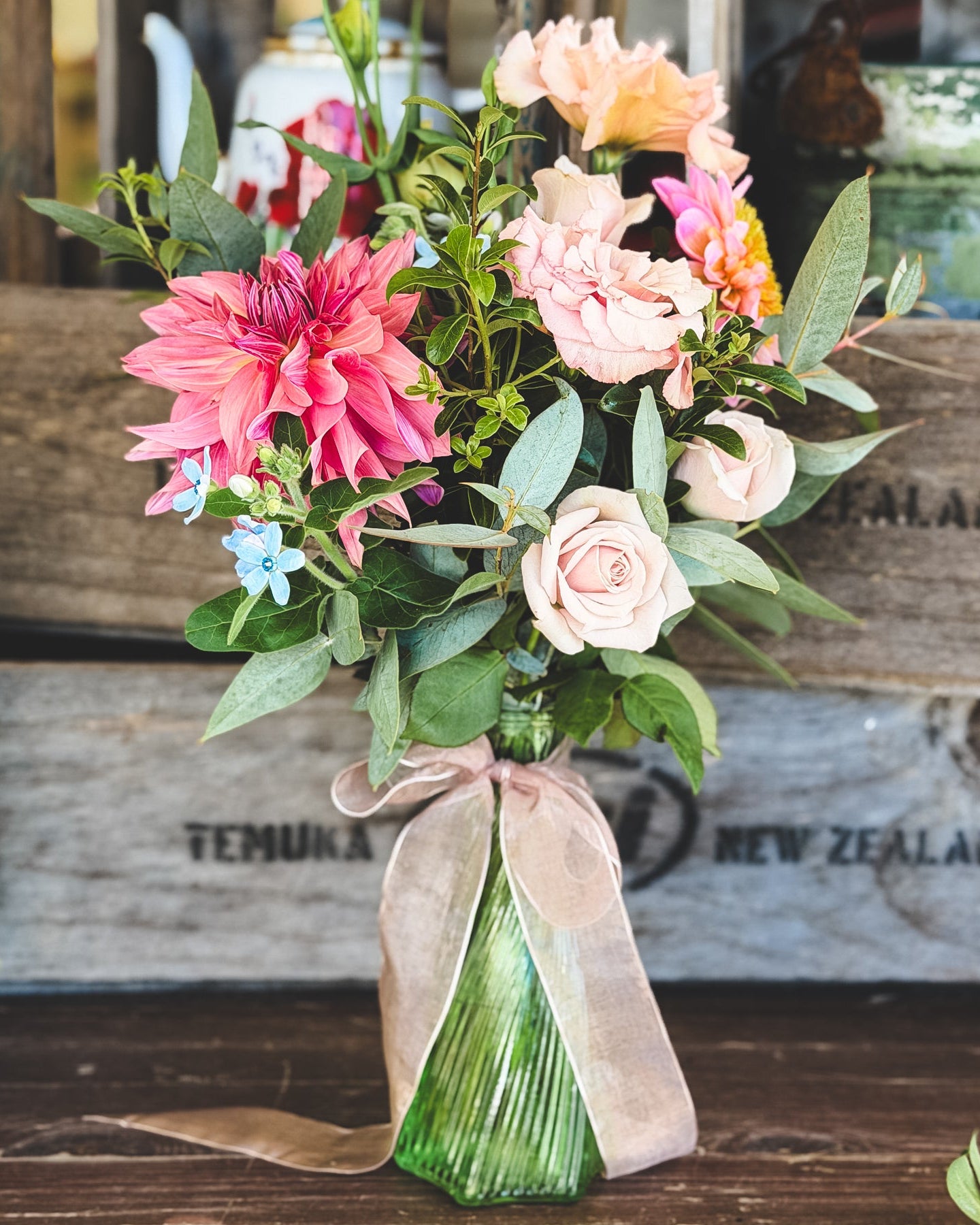 Bouquet of flowers with a green bud vase on a wooden surface, with a wooden crate in the background.