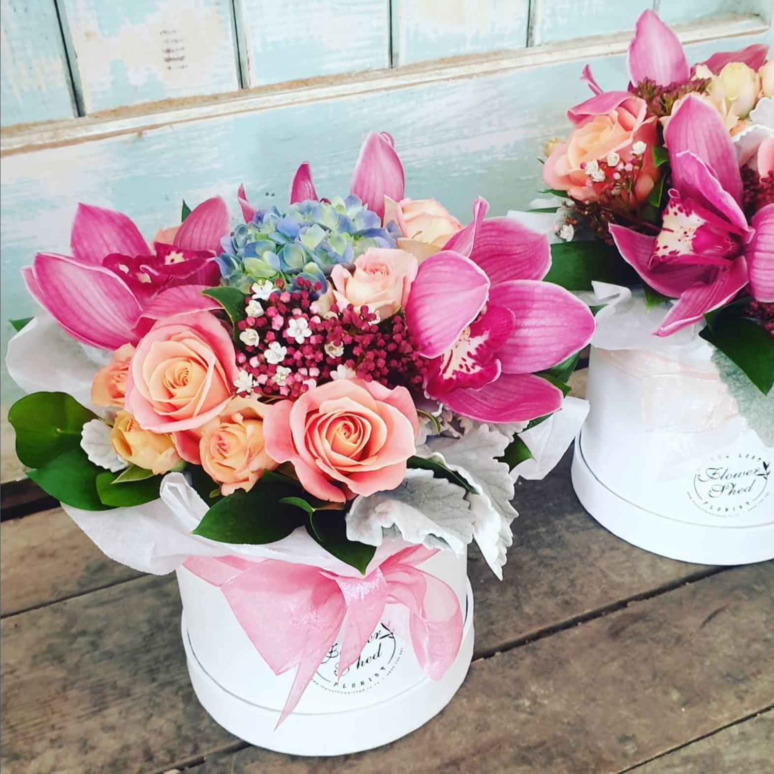 Two flower bouquets in white containers on a wooden surface with a light blue wall in the background.