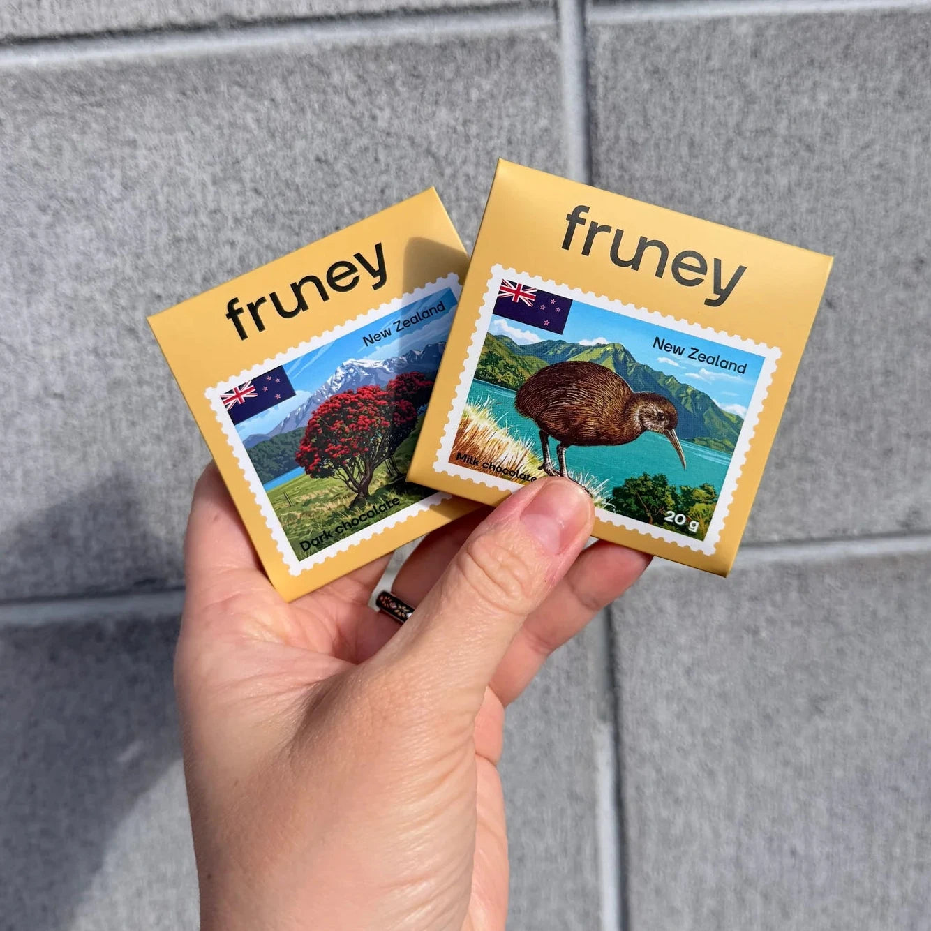 A hand holding two small square fruney magnets against a gray tiled wall. The magnets feature a yellow border with the word 'fruney' and a colorful landscape illustration of New Zealand.
