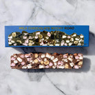 A milk chocolate rocky road bar with marshmallows and nuts, shown below its blue packaging box on a white marble background.