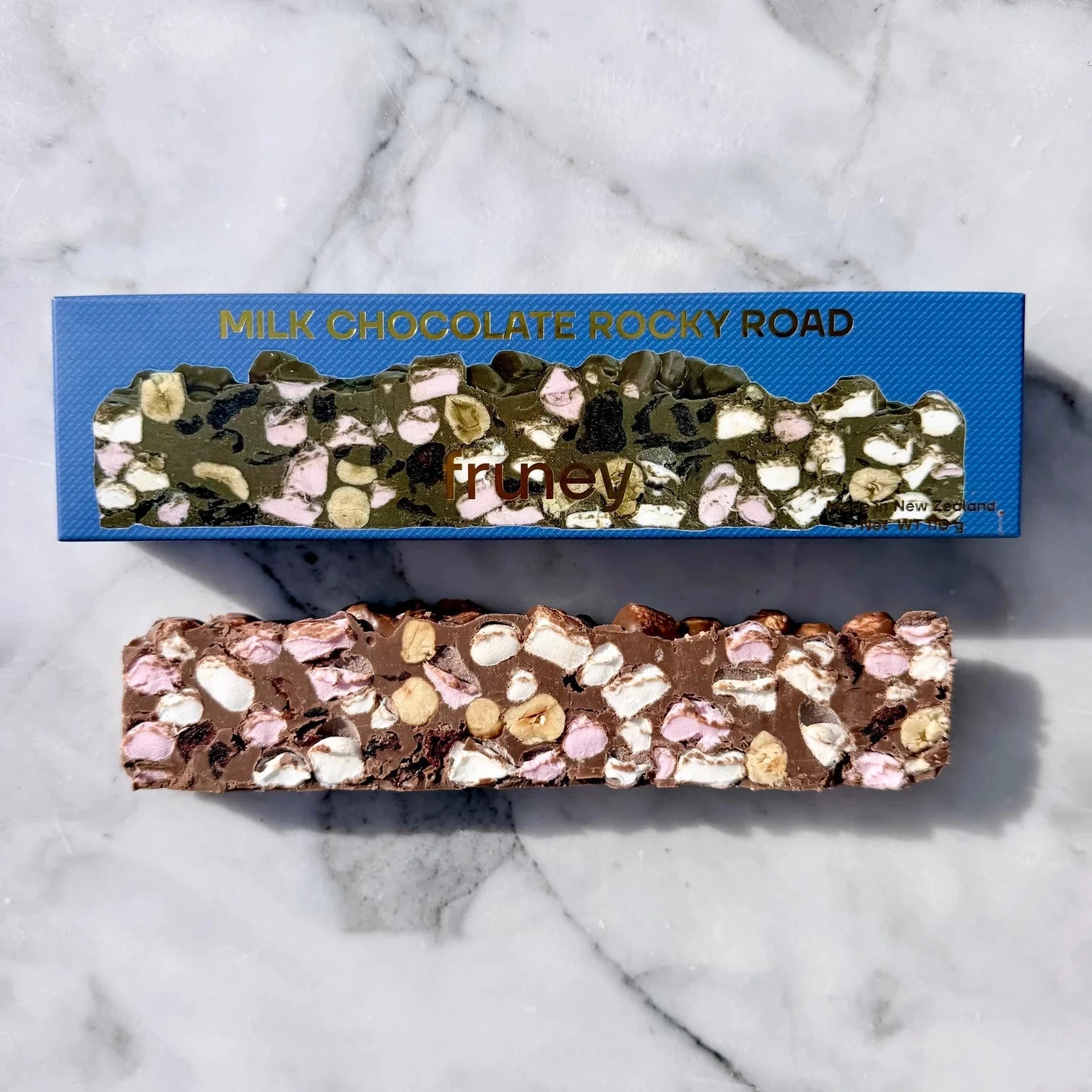 A milk chocolate rocky road bar with marshmallows and nuts, shown below its blue packaging box on a white marble background.