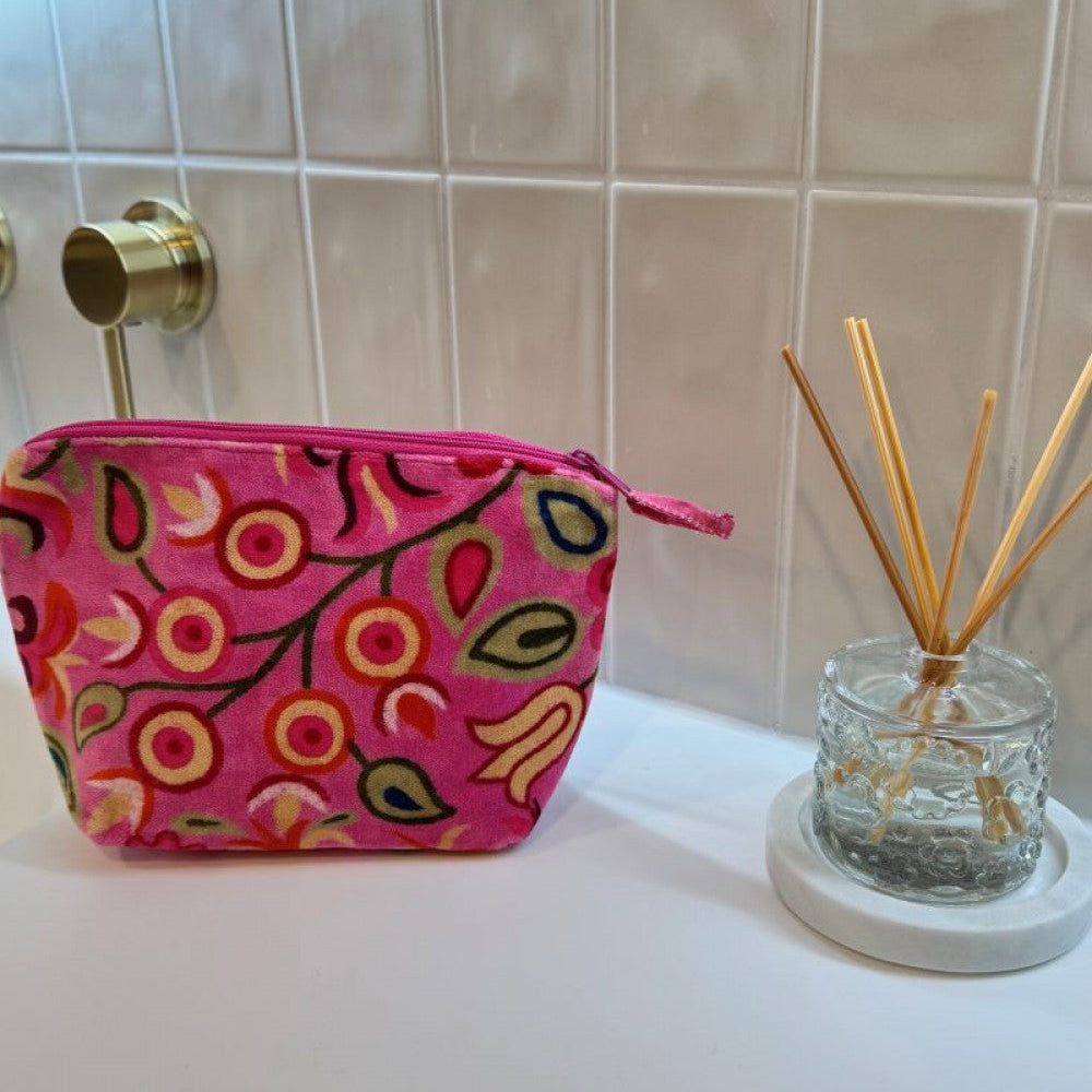 A pink zippered cosmetic pouch with a colorful floral and leaf pattern sits on a white countertop next to a glass reed diffuser against a beige tiled wall.